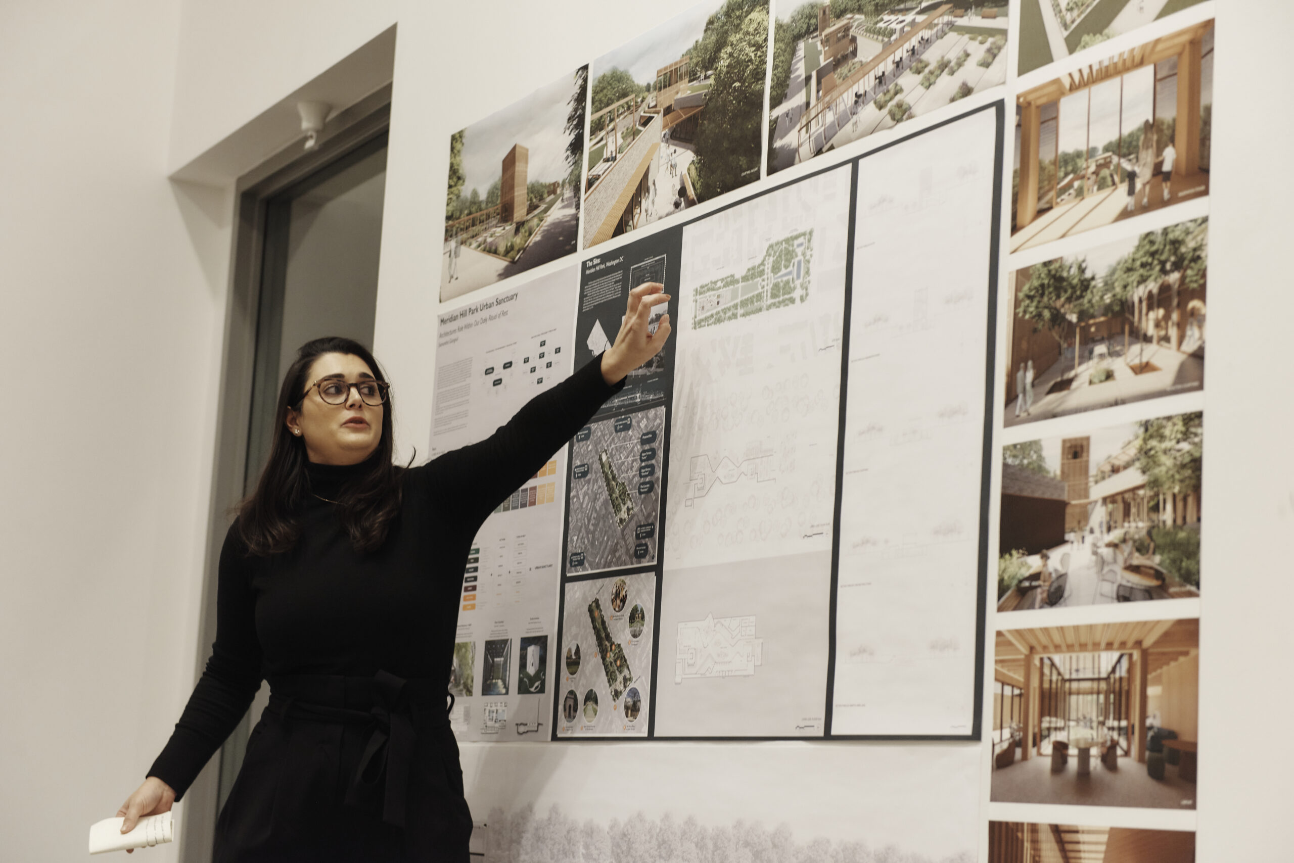 A woman in a black outfit and glasses gestures toward architectural design posters on a wall, showcasing building plans, renderings, and photographs. She seems to be explaining the designs, possibly highlighting various careers in architecture, in an indoor setting.