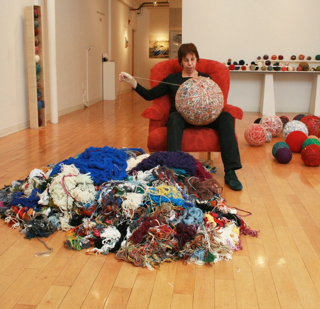 A person sits on a red chair in an art gallery, immersed in a month-long performance. They cradle a large, colorful ball of yarn as assorted yarn spills onto the floor. More vibrant yarn balls are displayed on tables behind them, marking a creative shift in time.