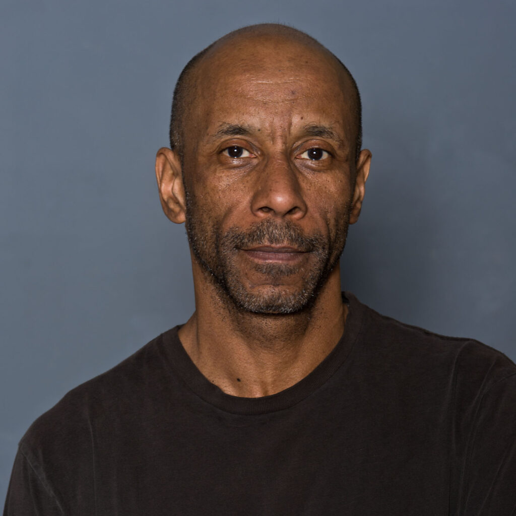 A man with a shaved head and a salt-and-pepper beard stands against a plain gray background, wearing a dark T-shirt. His neutral gaze meets the camera with the same focus as an undergraduate preparing for finals.
