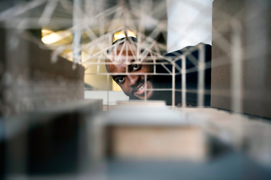 A friendly architect smiles and leans in to inspect a miniature architectural model, its detailed beams and structures clearly visible. The blurred background sharpens focus on both the model and the architects delighted expression.
