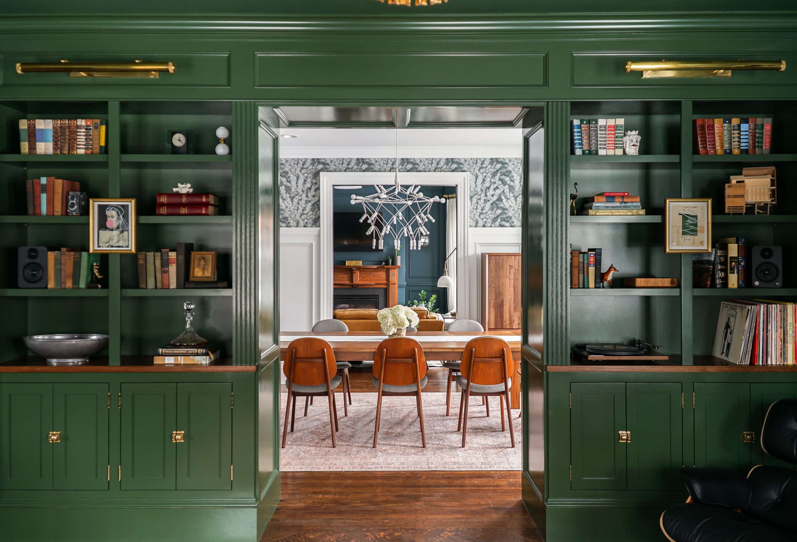 A sophisticated, renovated parlor with dark green walls and built-in bookshelves frames a view into the dining area. Shelves adorned with books, a trophy, and decor. The dining area features a modern chandelier and wooden furniture atop a patterned rug.