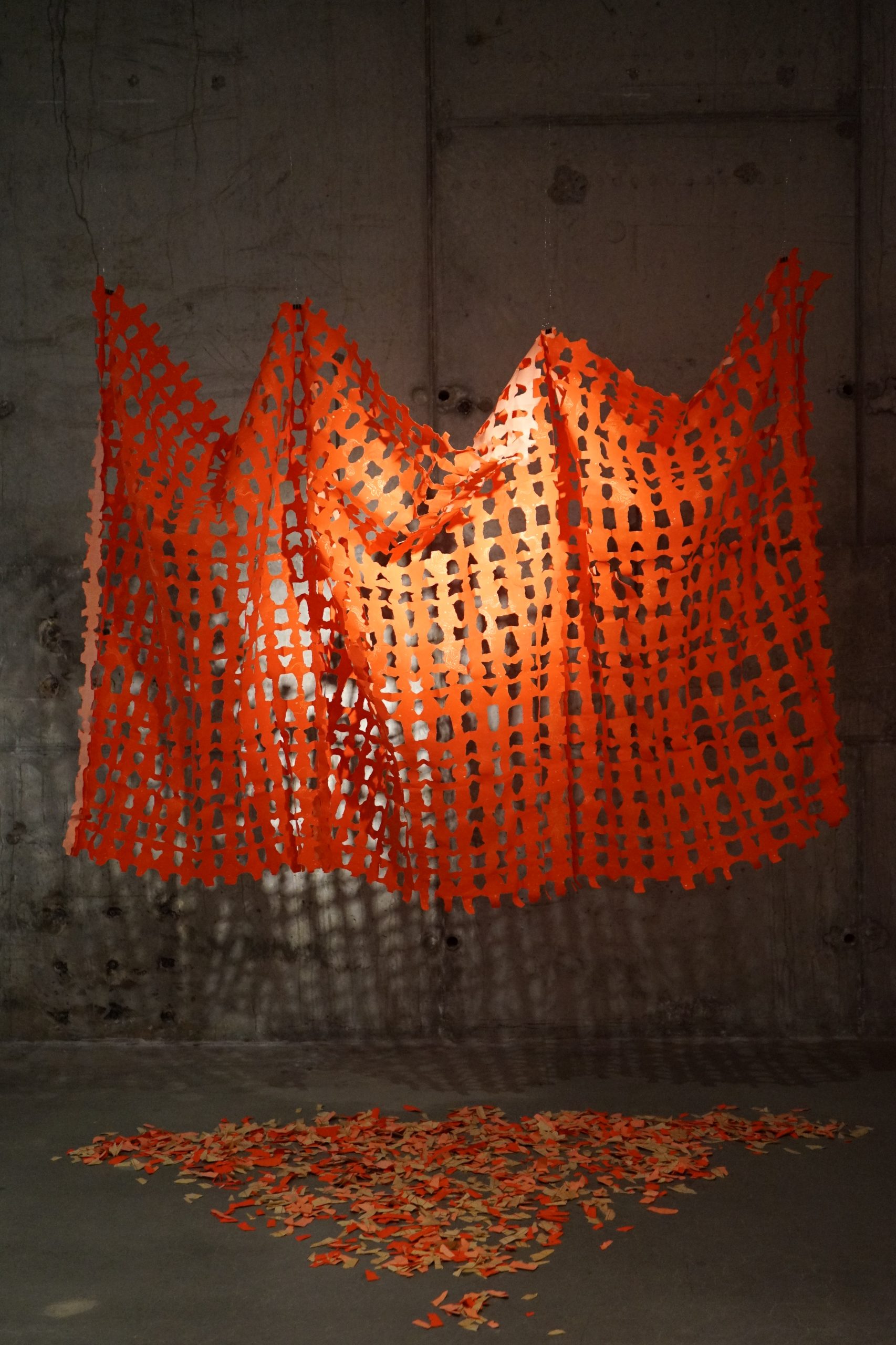 An orange construction fence drapes like fabric against a gray concrete wall. Small cut-out pieces are scattered below in an artful display. The light casts intricate shadows on the wall, enhancing the dramatic effect.