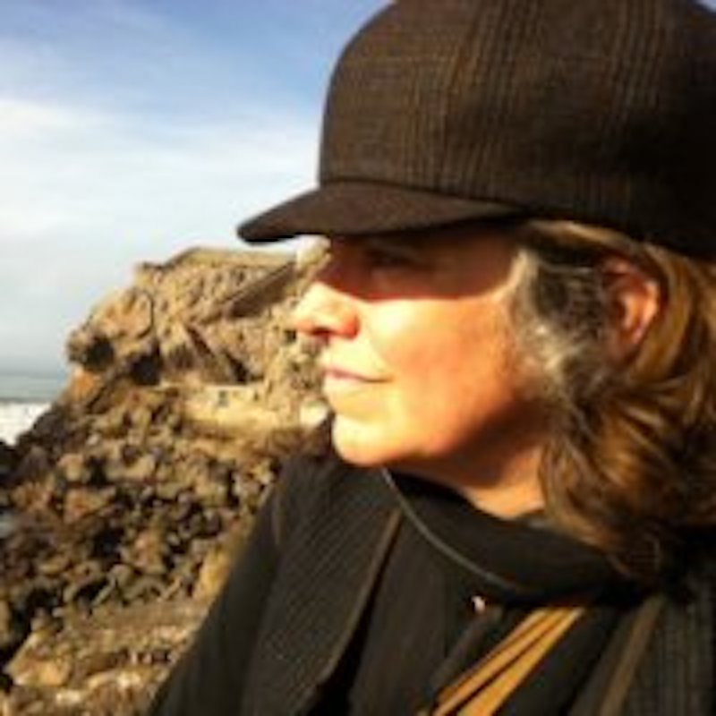 A person in a brown hat and scarf looks into the distance with rocky terrain and a bright sky in the background.