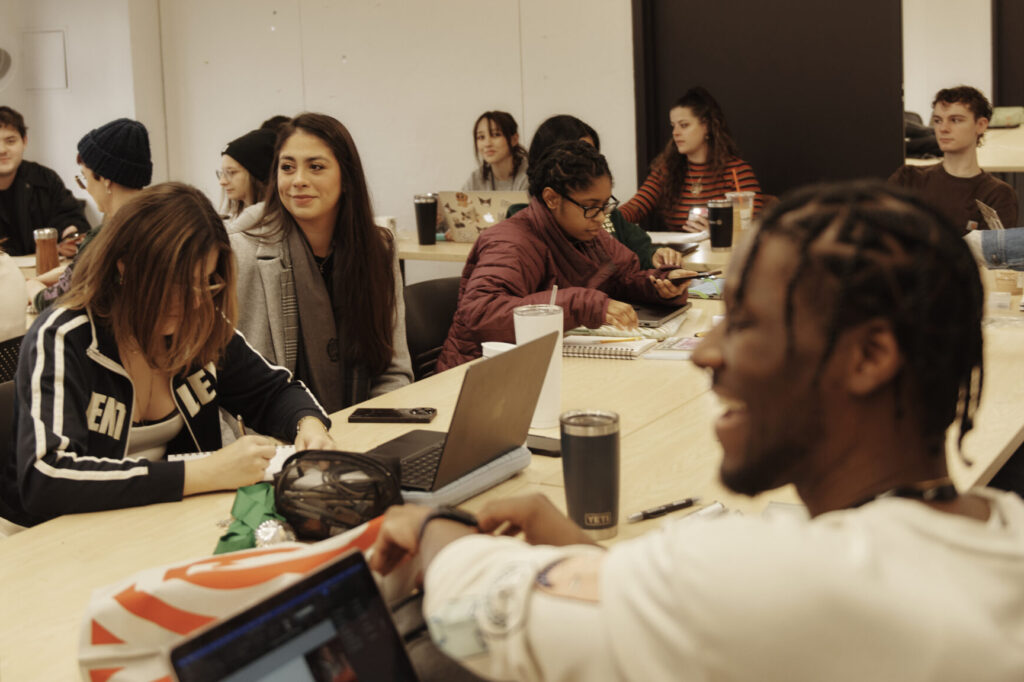 Students are gathered in a classroom, working and chatting around tables. Some have laptops open, while others write in notebooks. The atmosphere appears casual and collaborative.