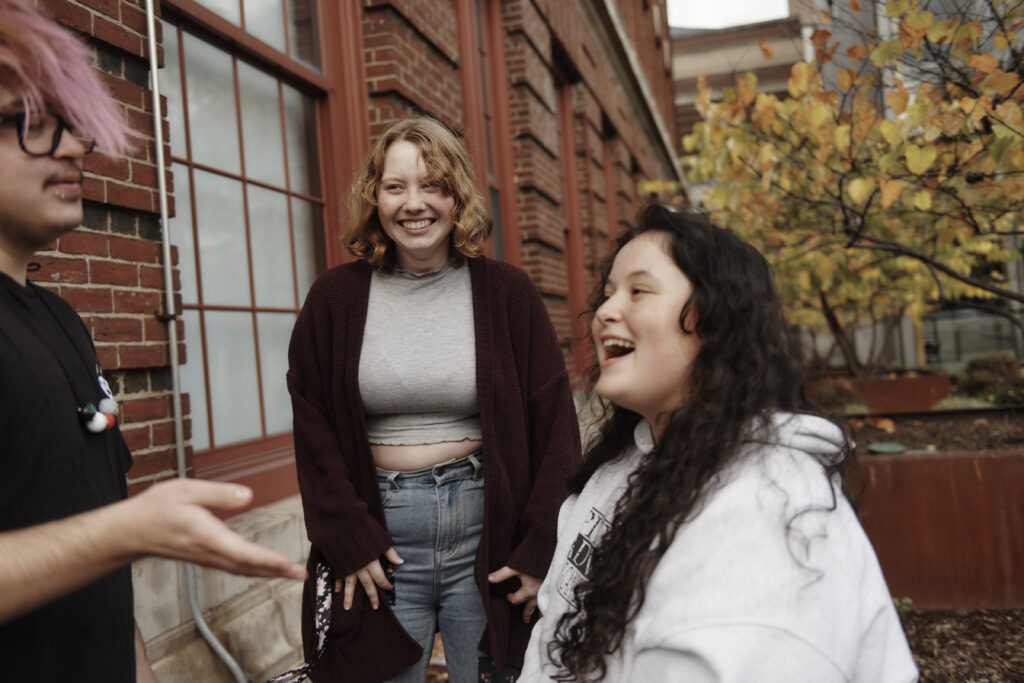 Three students smile and chat outside a brick building on MassArt's Campus. The person on the left has pink hair and gestures with their hand. The person in the middle wears a maroon cardigan and jeans, while the person on the right has long hair and a white hoodie.