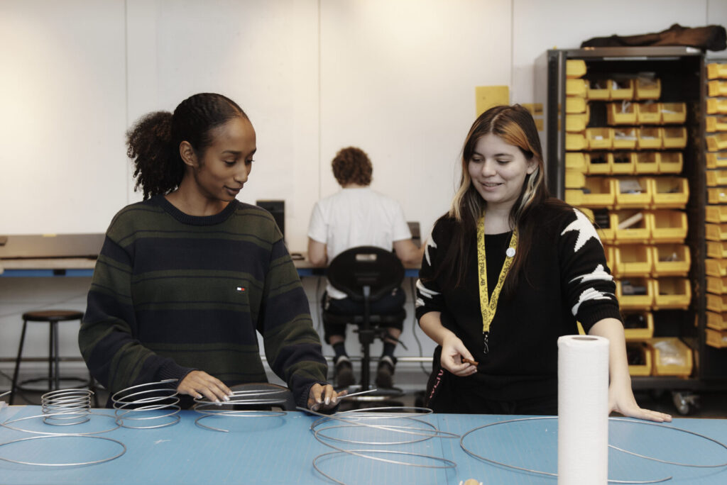Two women are standing at a table with metal coils, engaged in a project. They appear focused and are discussing the items. A person is seated at a desk in the background, facing away. Shelves with yellow bins line the wall.