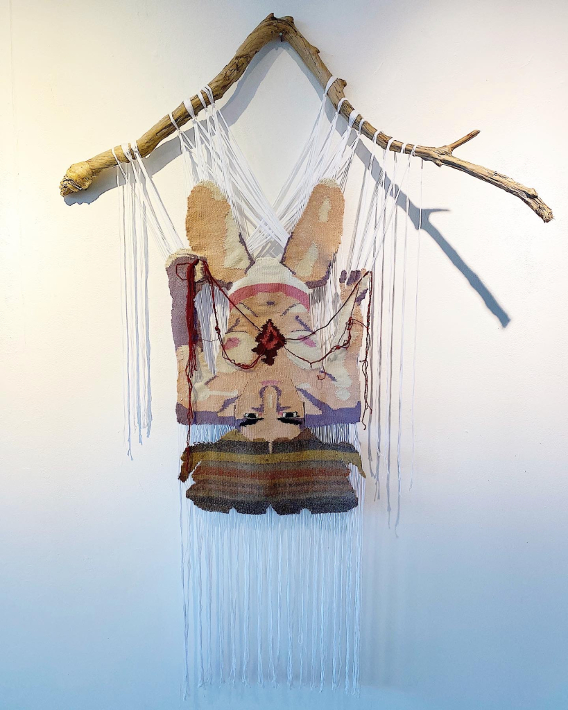 A woven tapestry of an upside-down human figure hangs from a natural wooden branch—a striking piece for your portfolio. The earthy colors, detailed facial features, and long yarn strands contrast beautifully against the plain white wall.