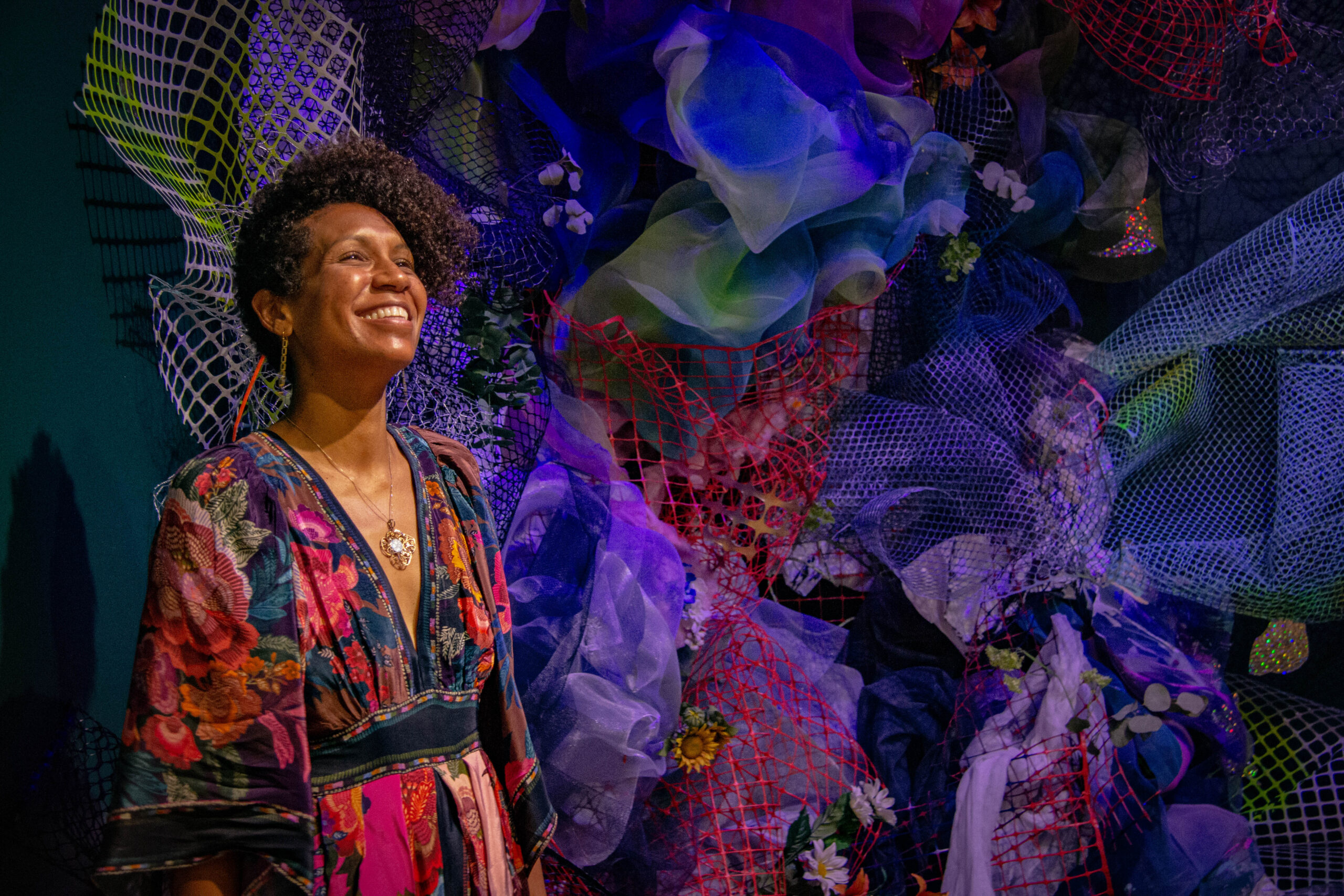 A person wearing a colorful floral dress beams with creative passion while standing beside an abstract art installation made of vibrant mesh, fabric, and netting. The background is a mix of purple, blue, and red hues, sparking wonder in a lively and artistic atmosphere.