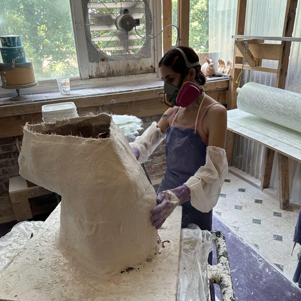 A person wearing protective gear, including a mask and gloves, is sculpting a large torso-shaped mold in a well-lit workshop. Various tools and materials are scattered around the workspace.