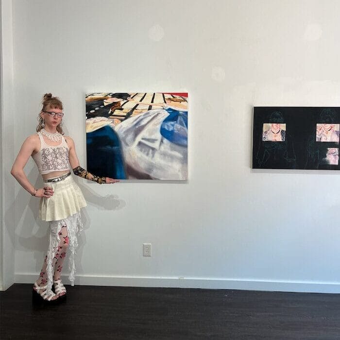 A person stands in an art gallery, posing next to a colorful abstract painting. They are wearing a white lace top, a cream skirt, and floral-patterned leggings. Another artwork is visible on the wall to the right.