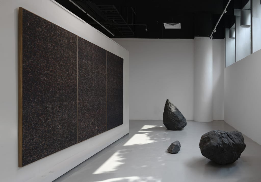 A minimalist art gallery features a large triptych with a dark, textured surface on the left wall. Three irregularly shaped rocks, including Batubuh #03, are placed on the light gray floor. Sunlight streams through upper windows, casting soft shadows.