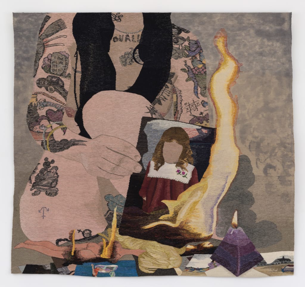 A detailed textile artwork depicting a person with numerous tattoos holding a burning photograph of a woman. Surrounded by small objects, including a purple pyramid, candles, and cigarette packs. The flames dramatically rise from the photo.