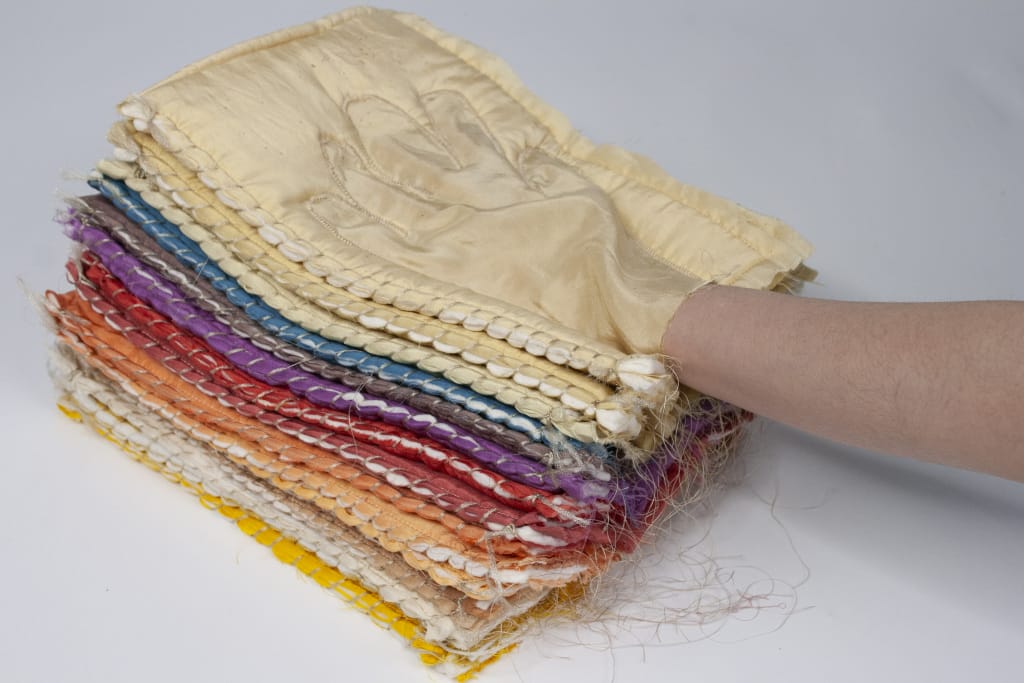 A hand wearing a beige glove reaches into a stack of textured fabric squares, layered in healing colors of yellow, orange, red, purple, blue, and cream against a light gray background.