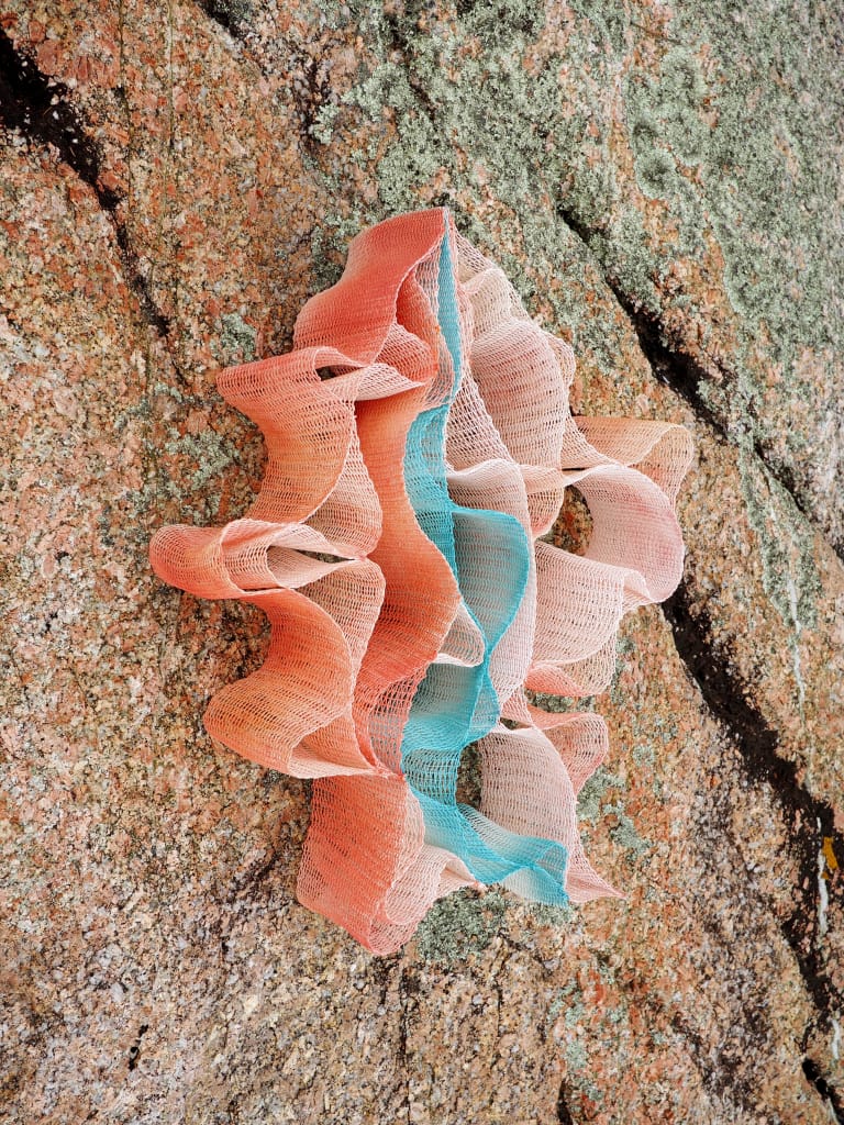 A multicolored mesh fabric, featuring shades of orange, blue, and white, is draped in wavy folds against a textured rock surface with patches of green and gray lichen.