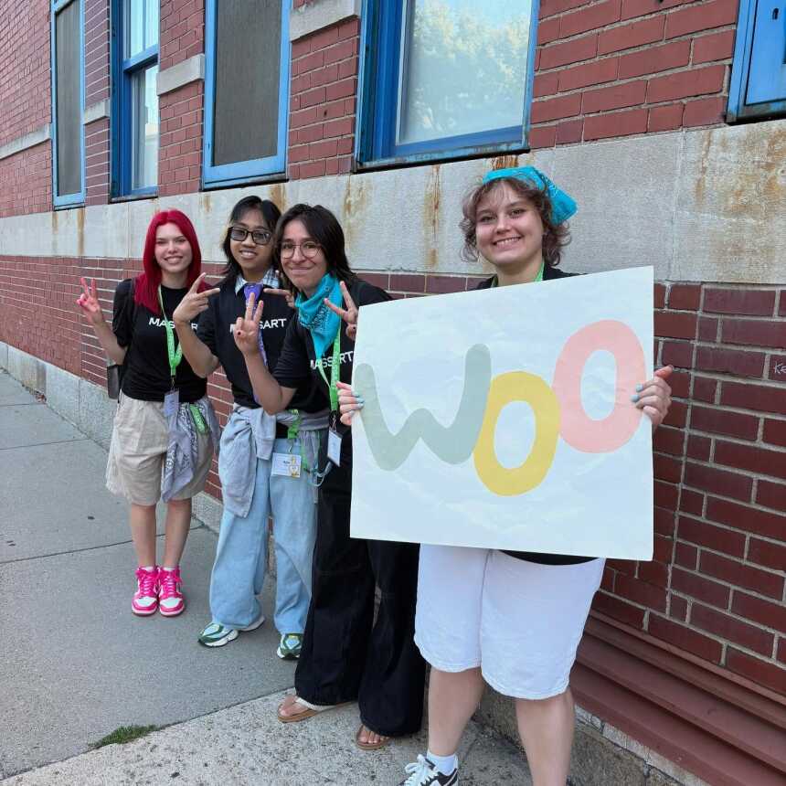 Four young people stand on a sidewalk by a brick building, smiling and making peace signs. One holds a colorful sign that says woo. They wear black shirts, name tags, and colorful accessories.