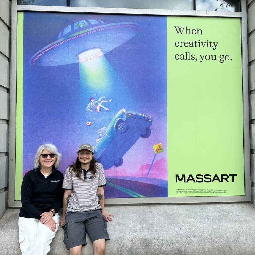 Two people sit smiling in front of a large MassArt poster showing a person and a car being beamed up by a UFO with the text, When creativity calls, you go. The background is a building with gray stone walls.