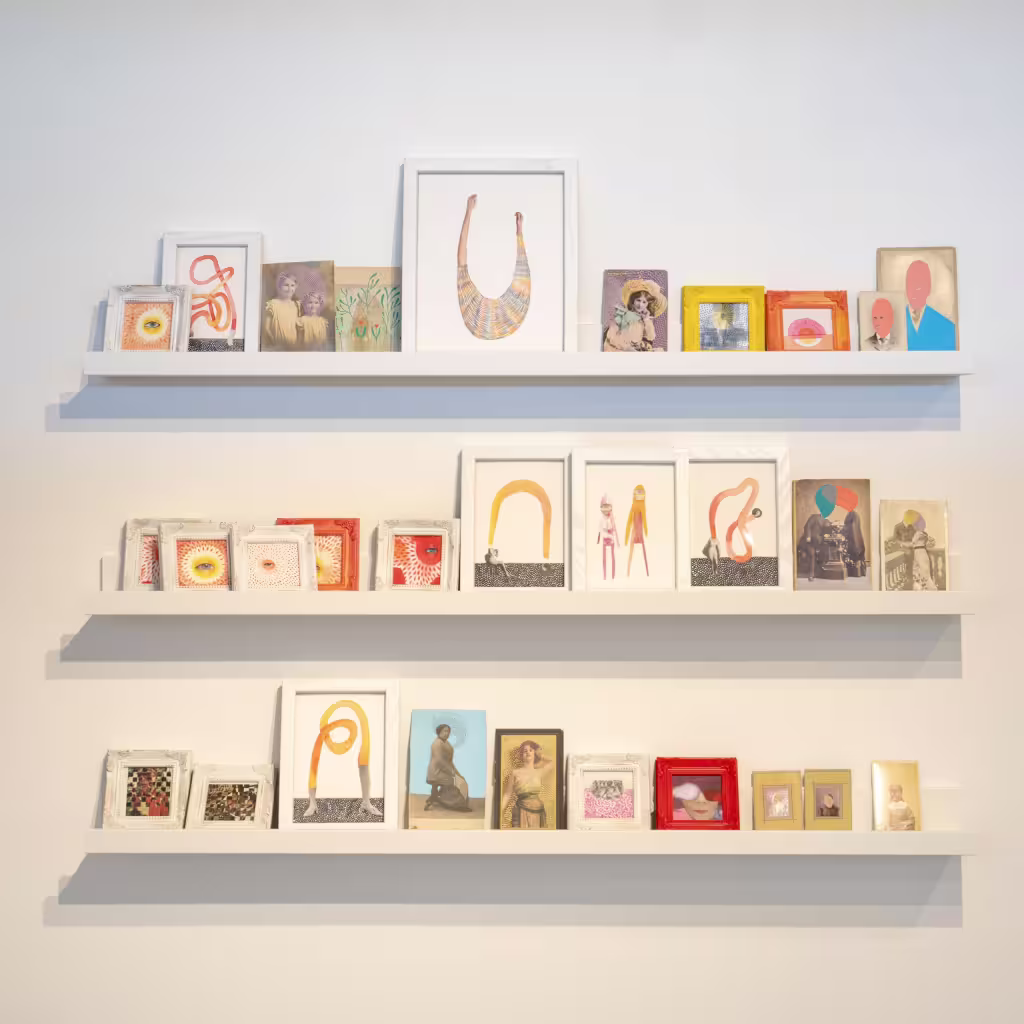 Three white shelves display small, colorful artworks and framed photos, reminiscent of MassArt exhibitions.